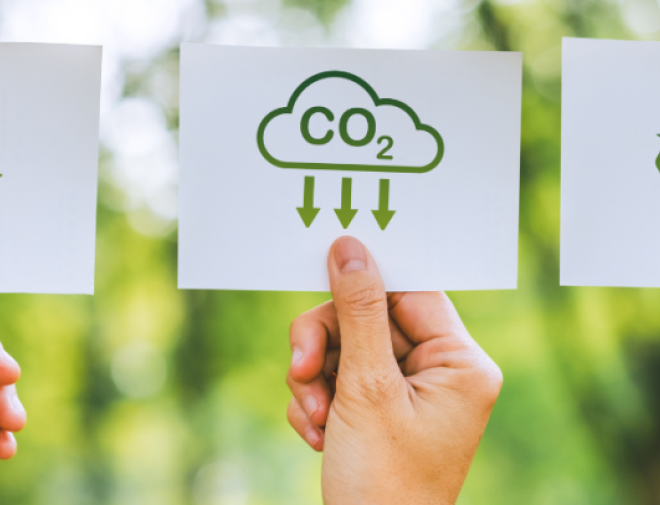 Three hands holding cards against a blurred green background. The cards display green environmental icons: on the left, a circular recycling symbol; in the middle, a cloud labeled “CO₂” with arrows pointing downward, representing carbon capture or reduction; and on the right, a recycling triangle symbol. This image conveys themes of sustainability and environmental responsibility.