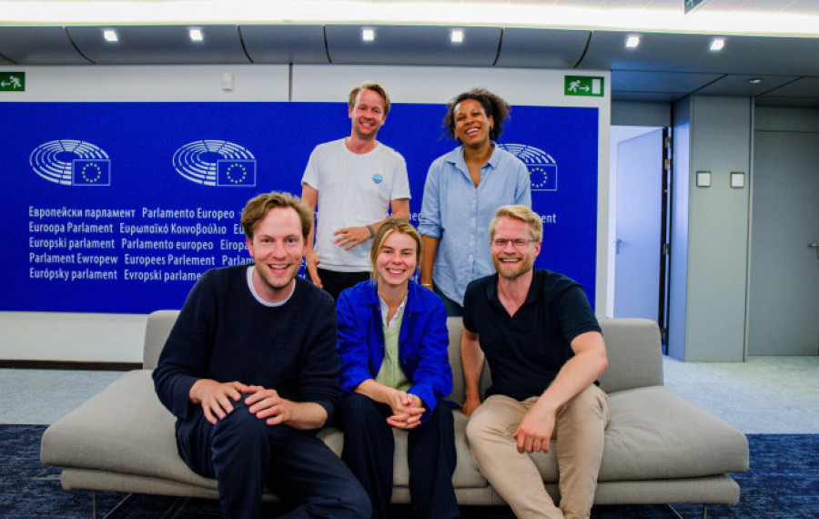 Group photo of five MEPs from Volt Europa at the European Parliament. They are seated on a light-colored couch in front of a blue wall with the European Parliament logo and multilingual text that reads “European Parliament.” From left to right: seated in front are Damian, Anna, and Kai, while Reinier and Nela stand in the back, all smiling. The setting is modern and well-lit.