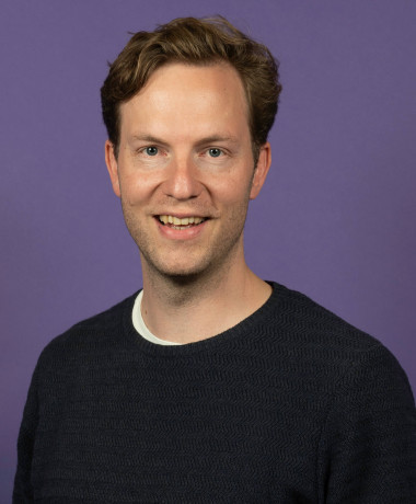 Portrait of Damian, smiling and wearing a dark sweater over a white shirt. He has short, wavy hair and stands against a solid purple background.