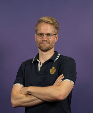 ortrait of Kai, standing with arms crossed and wearing a dark polo shirt with a small embroidered emblem. He has short blonde hair, glasses, and a slight smile, set against a solid purple background.